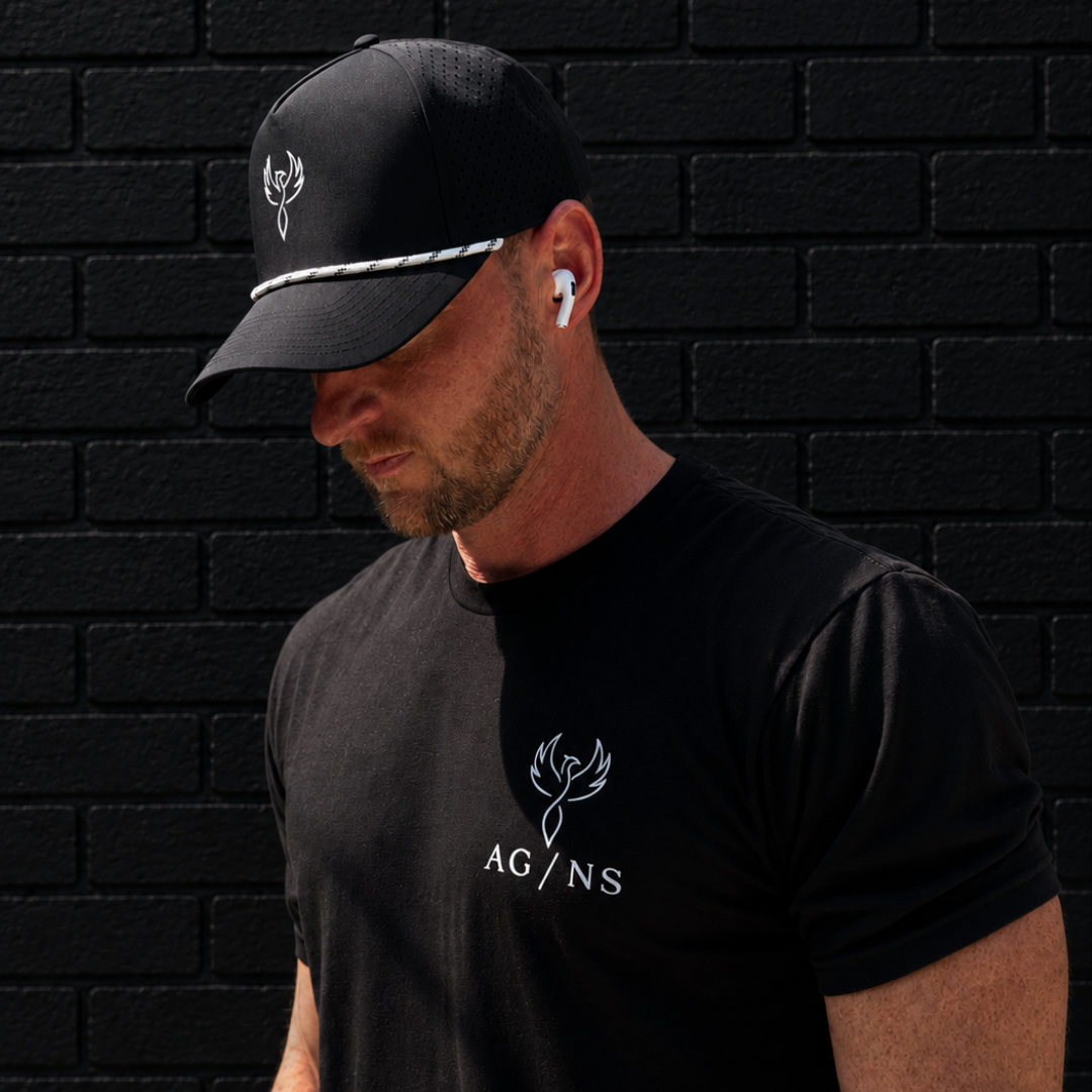 Man wearing a black cap and black t-shirt with a logo against a black brick wall.